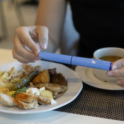 Close-up of hands holding an Ozempic injection pen while sitting at a table with a plate of food and a cup of soup.