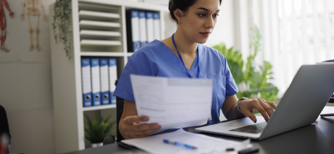 Focused young nurse or doctor in blue scrubs working on laptop while holding a medical report in modern office