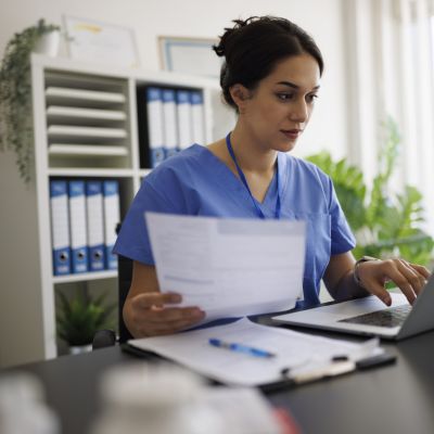 Focused young nurse or doctor in blue scrubs working on laptop while holding a medical report in modern office